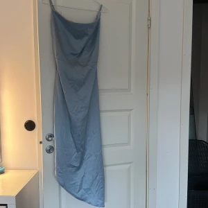 Ljusblå långklänning från Missguided - Säljer en ljusblå långklänning från Missguided i satinliknande material. Klänningen har smala axelband som går att justera, draperad halsringning och justerbart resårband längs ena sidan. Använd endast en gång på bröllop. Är i storlek 32