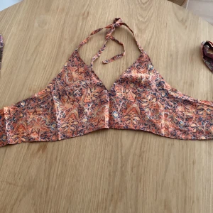 Blommig bikiniöverdel med knyt - Snygg bikiniöverdel med två olika sidor – ena sidan har ett färgglatt blommigt mönster i rosa, lila och grönt på brun botten, andra sidan har ett orange och rosa mönster. Knyt i nacken och ryggen för perfekt passform. Köpt utomlands 