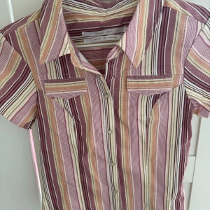 Randig skjorta  - Kortärmad skjorta från Urban Outfitters. Skjortan har smal passform, knappar framtill och två bröstfickor. Mönstret är randigt i rosa, vinrött, beige och vitt. Den är oanvänd och har lappen kvar. 