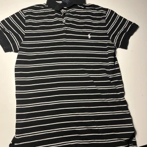 Randig pikétröja från Ralph Lauren - Svart och vit randig pikétröja från Polo Ralph Lauren i storlek S. Klassisk krage med knappar och broderad logga på bröstet. Tröjan är i mjuk bomull och har korta ärmar, perfekt för en clean och stilren look.