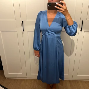 Blå omlottklänning från Neo Noir - Supersnygg blå långklänning från Neo Noir med omlottmodell och v-ringning. Klänningen har långa ärmar med lätt puff och är tillverkad i ett glansigt, mjukt material som ger en lyxig känsla. Perfekt för dig som vill sticka ut med stilren elegans.