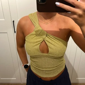 Glittrig guldig oneshoulder topp NA-KD - Säljer en glittrig guldig oneshoulder topp från NA-KD i storlek S. Toppen har en snygg twistad detalj vid axeln och en cut-out framtill. Materialet är stretchigt och skimrande, perfekt för att sticka ut. Passformen är tight och toppen är croppad.