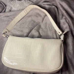 Vit axelväska i krokopräglat skinn - Snygg vit axelväska med croco-mönstrad yta i glansigt syntetskinn. Väskan har ett stilrent och minimalistiskt uttryck med silverfärgad dragkedja och matchande axelrem. Perfekt storlek för det viktigaste och ger en trendig touch till din outfit.