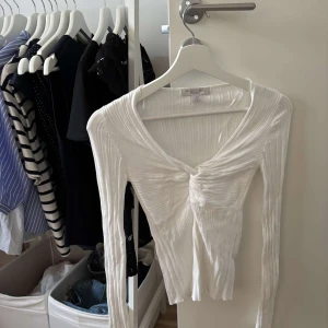Vit ribbad omlottopp - Supersnygg vit ribbad topp med omlottdesign och twistad detalj framtill. Långärmad och figurnära passform med v-ringning. Perfekt att styla med jeans eller kjol för en trendig look.
