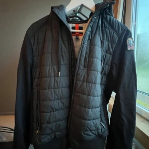 Svart quiltad jacka från Parajumpers - Snygg svart quiltad jacka från Parajumpers med huva och dragkedja. Jackan har ett stilrent rutmönster på framsidan, två fickor med dragkedja och en patch på ärmen. Använt ett fåtal gånger