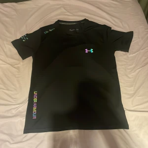 Svart Under Armour t-shirt - Svart t-shirt från Under Armour med färgglad logga på bröstet och text längs sidan. Klassisk rund hals och korta ärmar. Perfekt för dig som gillar sportig och clean stil. Passar både M och S