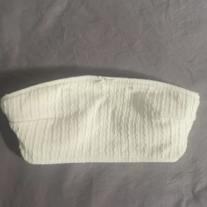 Vit stickad bandeau tubtopp med rosett - Supersöt vit bandeau tubtopp med stickat kabelmönster och liten rosett framtill. Toppen är fodrad på insidan för extra komfort och har en mjuk, elastisk känsla. Perfekt att styla med jeans eller kjol för en trendig look.