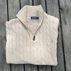 Beige kabelstickad half zip från Polo Ralph Lauren - Kabelstickad beige tröja från Polo Ralph Lauren med half zip och hög krage. Klassisk broderad logga på bröstet och ribbade muddar. Perfekt till jeans för en clean och stilren look. Storlek L men passar mer som M/ 175-185 cm