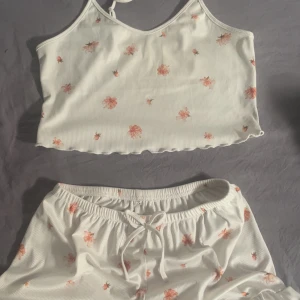 Vit blommig croppad linnetopp och shorts - Supersöt croppad linnetopp i vit ribbad bomull med små rosa blommor och shorts. Toppen har smala axelband och vågig kant nertill för en extra gullig touch. Perfekt för varma dagar eller som mysig pyjamasöverdel. Matchar med shorts i samma mönster.