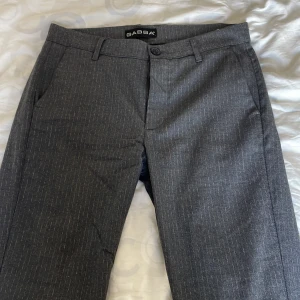 Gabba grå kostymbyxor med pinstripe - Snygga grå kostymbyxor från Gabba med diskret pinstripe-mönster. Klassisk passform med hällor för bälte och knappstängning. Perfekta för en clean och stilren look. Tillverkade i ett bekvämt material som funkar året runt.