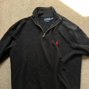 Marinblå half zip tröja från Polo Ralph Lauren - Blå långärmad tröja från Polo Ralph Lauren med half zip och klassisk röd broderad logga på bröstet. Tröjan har en hög krage och är tillverkad i bomull med en lätt stickad struktur. Perfekt för dig som gillar stilren och tidlös design.