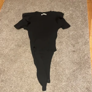 Svart bodytopp med volangärm från Zara - Svart bodytopp från Zara med korta ärmar och snygga volangdetaljer vid axlarna. Rund halsringning och figurnära passform. Perfekt att styla med jeans eller kjol för en clean look.