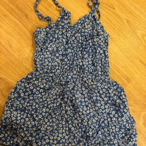 Blå blommig byxdress H&M Divided - Supersöt blå byxdress från H&M Divided med vitt blommönster. Dressen har smala axelband och markerad midja, perfekt för varma dagar. Lätt och luftig känsla, passar dig som gillar en avslappnad men ändå söt stil.