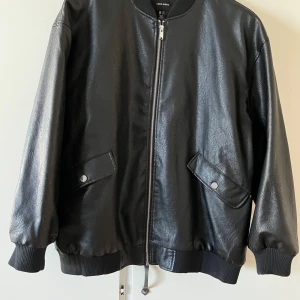 Svart bomberjacka i skinn från Vero Moda - Snygg svart bomberjacka från Vero Moda i skinnimitation. Jackan har dragkedja framtill, två fickor med tryckknappar och ribbade muddar vid ärmslut och nederkant. Perfekt för dig som vill ha en stilren och trendig look.
