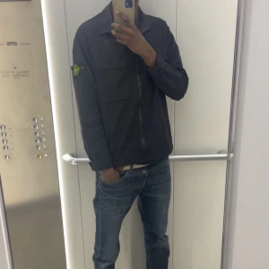 Svart overshirt från Stone Island - Svart overshirt från Stone Island med klassisk krage, två stora bröstfickor och dragkedja framtill. Jackan har den ikoniska Stone Island-loggan på ärmen och är tillverkad i ett lätt syntetmaterial. Perfekt för lager-på-lager och stilren streetwear. Jackan passar på folk mellan 175-185. För bilder när jag har på mig den skriv pm🤗