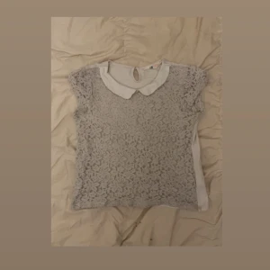 Vit spetsblus med krage - Supersöt vit blus med spets framtill och klassisk rund krage. Framsidan har ett detaljerat blommigt spetsmönster och ärmarna är korta. Baksidan är slät i mjukt tyg och har en liten knapp i nacken. Perfekt för dig som gillar romantisk och feminin stil.