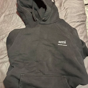 Svart hoodie från Ami Paris - Svart hoodie från Ami Paris med vit broderad logga på bröstet. Klassisk modell med huva, snörning och stor magficka framtill. Mjuk bomullskvalitet och ribbade muddar vid ärmslut och nederkant. Perfekt för en chill och stilren look.