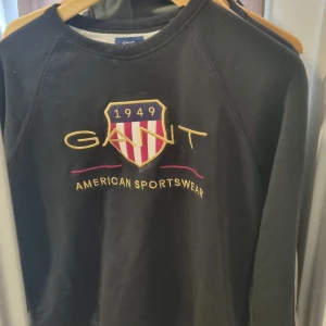 Svart sweatshirt från GANT med brodyr - Svart sweatshirt från GANT med broderad logga och sköld på bröstet. Tröjan har rund halsringning, långa ärmar och ribbade muddar. Snygg kontrast med gult och rött broderi samt texten 'American Sportswear'. Perfekt för en sportig och stilren look.