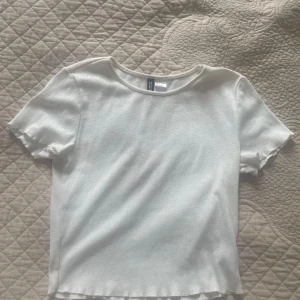 Vit ribbad topp från H&M Divided - En stilren vit ribbad topp från H&M Divided i storlek S. Toppen har korta ärmar, rund halsringning och diskret struktur i tyget. Perfekt att matcha med jeans eller kjol för en clean look.