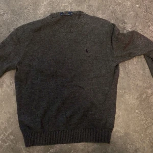 Mörkgrå stickad tröja Polo Ralph Lauren - Mörkgrå stickad tröja från Polo Ralph Lauren med diskret broderad logga på bröstet. Klassisk rund halsringning och ribbade muddar vid ärmslut och nederkant. Perfekt för lager-på-lager under kyliga dagar.
