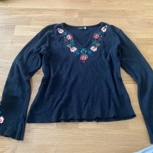 Svart broderad  tröja - Svart stickad tröja från Add it med v-ringning och färgglada broderade blommor runt halsen och på ärmarna. Långärmad modell med mjukt material och lätt utsvängd nederkant. Storlek M men funkar för S oxå