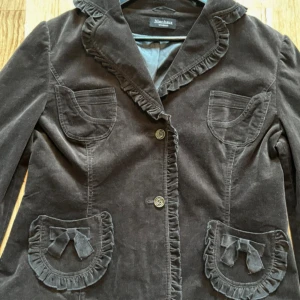 Brun manchesterkavaj med volangdetaljer - Snygg brun kavaj i manchester från Nienhaus Woman. Jackan har volangkant längs kragen och fickorna, samt söta rosetter på de nedre fickorna. Två knappar framtill och fyra fickor ger en cool och unik look. Perfekt för dig som gillar detaljer och vintagevibbar.