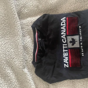 Svart t-shirt Zavetti Canada med storleken L i barn storlekar - Svart t-shirt från Zavetti Canada med tryck framtill i vitt och rött samt en broderad lönnlövssymbol. Klassisk rund halsringning med röd och vit detalj på insidan av kragen. Skön passform och korta ärmar, perfekt till jeans eller shorts.