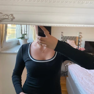 Svart långärmad topp med vit kant - Snygg svart långärmad topp med en bred, rundad urringning och vit kant runt halsen. Tight passform som framhäver figuren och ger en stilren look. Perfekt att matcha med jeans eller kjol för en clean och trendig stil. Använd 1 gång.