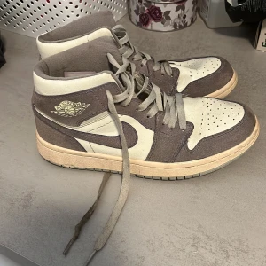 Nike Air Jordan 1 i beige och grå mocka - Säljer ett par Nike Air Jordan 1 sneakers i beige och grå färg. Skorna har klassisk hög siluett, snörning och ikonisk Air Jordan-logga på sidan. Ovandelen är i en mix av mocka och skinn, med rund tå och platt sula. Perfekta för dig som gillar streetstyle.