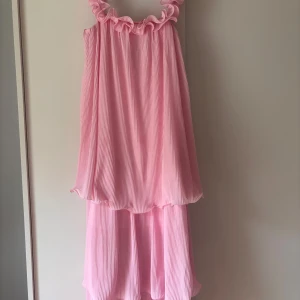 Ljusrosa volangklänning med öppen rygg - Superfin ljusrosa långklänning med volangkant vid axlarna och plisserat tyg. Klänningen har öppen rygg med knytband och dubbla lager för extra flow. Perfekt för sommarens alla tillfällen när du vill känna dig extra söt och bekväm.  Jag är 160 och klänningen går ner över fötterna. Där av lite slitage längst ned på klänningen men inget som syns tydligt när den är på! Klänningen är ganska stor i storlek då jag vanligtvis är en S men XS passade bra