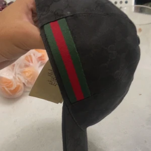 Gucci keps - Snygg svart keps från Gucci med diskret GG-monogram i tyget och klassisk röd och grön rand på sidan. Kepsen har böjd skärm och är tillverkad i ett slitstarkt material som känns lyxigt. Perfekt för dig som vill ha en stilren accessoar med exklusiv känsla.
