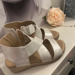 Silvriga sandaler från Zoey, strl 34 - Snygga sandaler i silverfärgat material från Zoey. De har breda remmar, dragkedja bak och en platt sula med repdetalj runt kanten. Barnskor storlek 34. Köpte för 350kr säljer för 65kr 