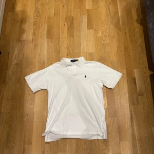 Vit pikétröja från Ralph Lauren - Klassisk vit pikétröja från Ralph Lauren med korta ärmar och broderad marinblå logga på bröstet. Tröjan har krage, knappar vid halsen och är gjord i mjuk bomull. Perfekt för en clean och stilren look.Ny skick! Nypris runt 1000