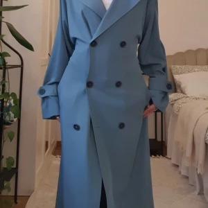 Blå trenchcoat med bälte och knappar - Snygg blå trenchcoat med dubbelknäppning och bälte i midjan. Jackan har långa ärmar med knappar vid ärmslut och klassisk krage. Perfekt för dig som vill ha en stilren och trendig look till vardags. Lång modell som ger en cool siluett.