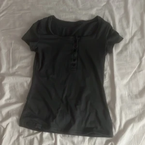 Svart topp med knappar fram - En svart, enkel topp med korta ärmar och rund halsringning. Framsidan har en knappslå med flera svarta knappar som ger en snygg detalj. Perfekt till jeans eller kjol för en clean look.