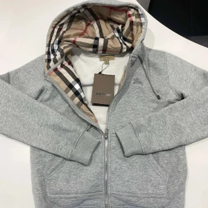 Grå Burberry hoodie med rutigt foder - Snygg grå hoodie från Burberry med dragkedja och klassiskt rutigt foder i huvan. Tröjan har långa ärmar, känguruficka och ribbade muddar. Perfekt för dig som vill ha en stilren och lyxig vardagslook.