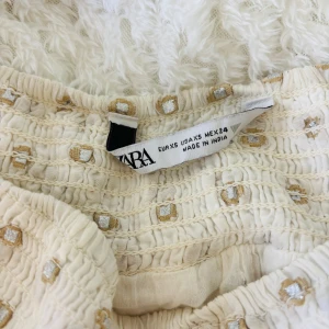 Zara kjol - Supersöt kort volangkjol från Zara i beige/vit med guldiga och vita broderade detaljer. Kjolen har smock i midjan för extra komfort och en luftig, flowy känsla. Perfekt för dig som gillar en feminin och trendig stil.