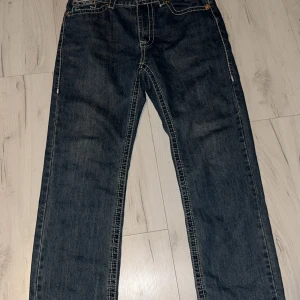 True Religion jeans med kontrastsömmar - Säljer ett par klassiska blå jeans från True Religion med tydliga vita kontrastsömmar och ikoniska bakfickor. Jeansen har normal passform och raka ben, samt detaljerade knappar och dragkedja. Perfekta för dig som gillar streetwear och snygga detaljer.