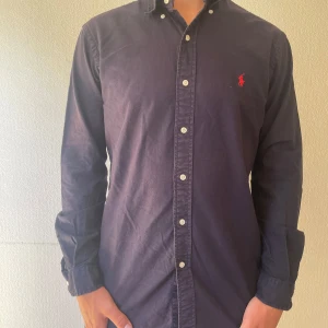 Mörkblå skjorta från Ralph Lauren - Säljer en mörkblå skjorta från Ralph Lauren i custom fit. Skjortan har klassisk krage, knäppning framtill och det ikoniska röda broderade logot på bröstet. Tillverkad i bomull och har långa ärmar. Perfekt för dig som gillar stilrena och tidlösa plagg.