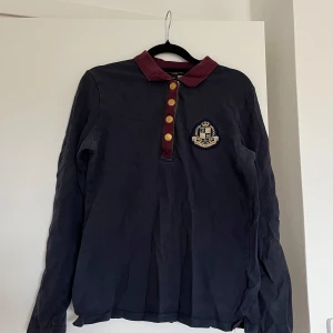Mörkblå pikétröja JH collection  - Snygg mörkblå långärmad pikétröja från JH Collection med vinröd krage och knappslå. Guldiga knappar och broderat emblem på bröstet.