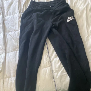 Svarta mjukisbyxor från Nike - Svarta mjukisbyxor från Nike med broderad vit logga på benet. Byxorna har resår i midjan och dragsko för skön passform. Perfekta för chill eller träning, med klassisk stil och mjukt material. Skriv till mig för frågor, pris kan diskuteras😃