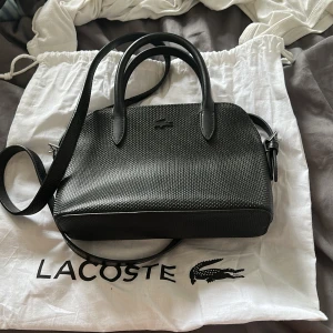 Svart handväska från Lacoste - Snygg svart handväska från Lacoste med stilren struktur och diskret krokodillogga framtill. Väskan har både handtag och justerbar axelrem samt dragkedja upptill. Perfekt storlek för det viktigaste och en clean look till alla outfits. Använd två gånger, dustbag ingår.