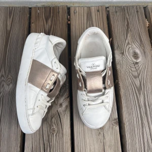 Valentino Garavani open sneakers - Snygga vita Valentino Garavani Rockstud sneakers i skinn med metallic guldbrun detalj över snörningen. Klassisk rund tå, platt sula och ikoniska nitar baktill. Perfekta för dig som vill ha en lyxig och trendig look.