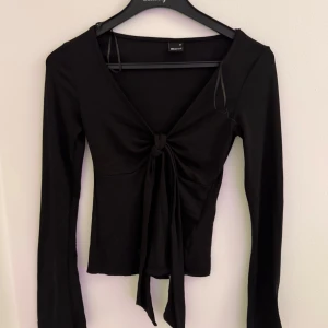 Svart långärmad topp med knyt - Snygg svart långärmad topp från Weekday med djup v-ringning och knyt framtill. Modellen är slim och har en croppad känsla, perfekt att styla med höga jeans eller kjol. Enkel men trendig design som passar till många olika looks.