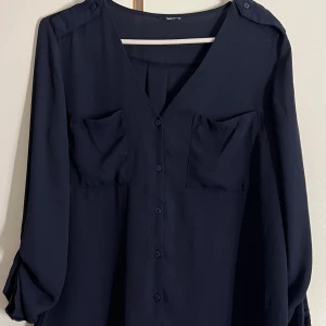 Mörkblå blus med v-ringning och knappar - Snygg mörkblå blus med v-ringning, knappar framtill och två bröstfickor. Långärmad modell med lite lösare passform och axelremmar med knappdetalj. Perfekt till jeans eller kjol för en clean look.