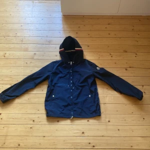 Blå vindjacka från Moncler med huva - Snygg marinblå vindjacka från Moncler med huva och dragkedja framtill. Jackan har Moncler-logga på ärmen och vita knappar vid fickorna. Huvan har en stilren randig detalj. Perfekt för dig som vill ha en clean och sportig look.