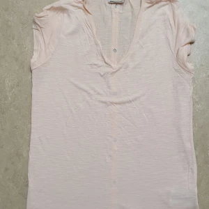 T-shirt - Supersöt ljusrosa t-shirt i tunn bomull med v-ringning och knappar hela vägen framtill. Kortärmad och perfekt att styla med jeans eller kjol. Enkel och clean look som funkar till många outfits.