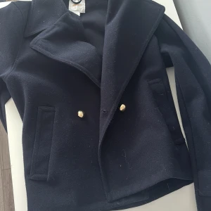 Mörkblå kappa från Gina Tricot - Stilren mörkblå kappa från Gina Tricot med snygga guldknappar framtill och klassisk krage. Jackan har två fickor och är tillverkad i ett ulligt material som ger en lyxig känsla. Perfekt för dig som vill ha en tidlös look med edge.