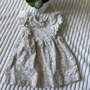 Blommig kortklänning från Newbie 68, kids - Supersöt kortklänning från Newbie med blommigt mönster i pastellfärger. Klänningen har volangprydda ärmlösa axlar med spetsdetaljer och är gjord i mjuk bomull. Perfekt för sommarens alla tillfällen!