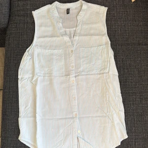 Randig ärmlös skjorta från H&M - Snygg ärmlös skjorta från H&M i ljusblått och vitt med tunna ränder. Skjortan har två bröstfickor, knappar framtill och en avslappnad passform. Perfekt för varma dagar och enkel att styla till jeans eller shorts.